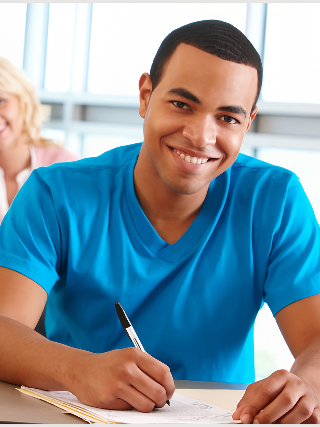 A smiling student writes at a desk in class.