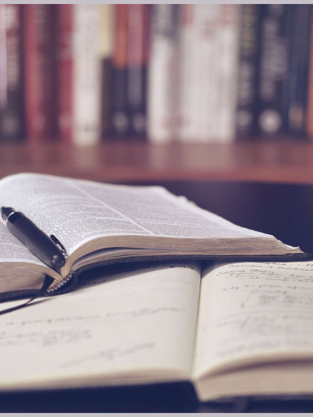 Open books, notes, and a pen sit in front of a bookshelf.