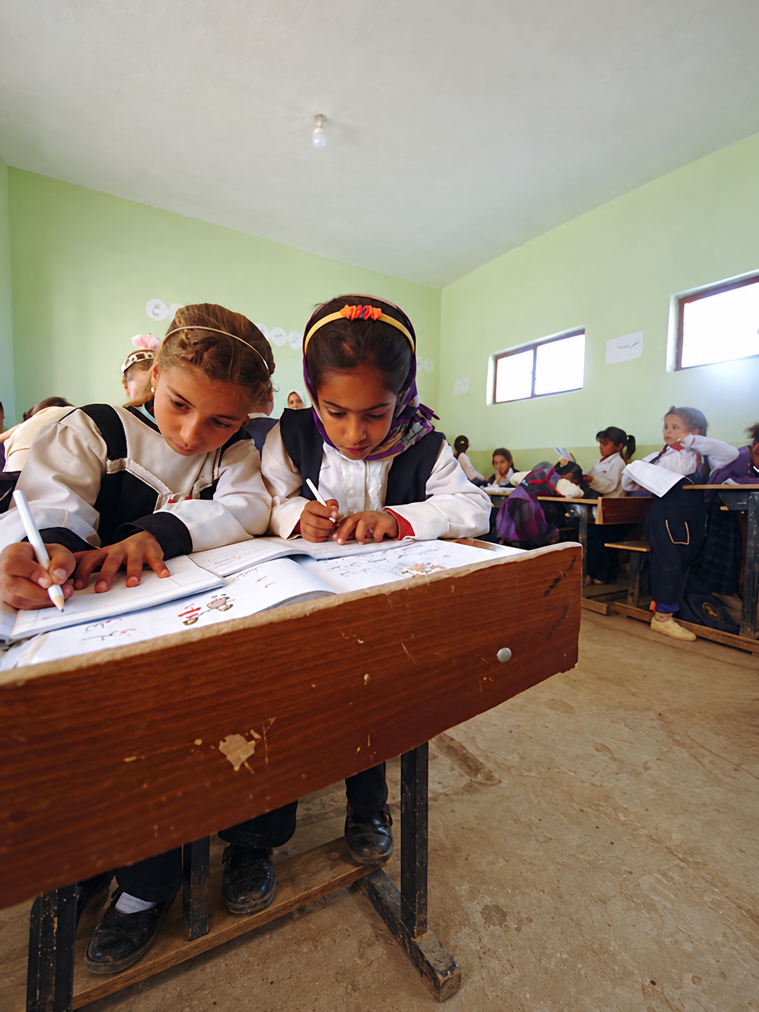 Two schoolgirls work together at a classroom desk.