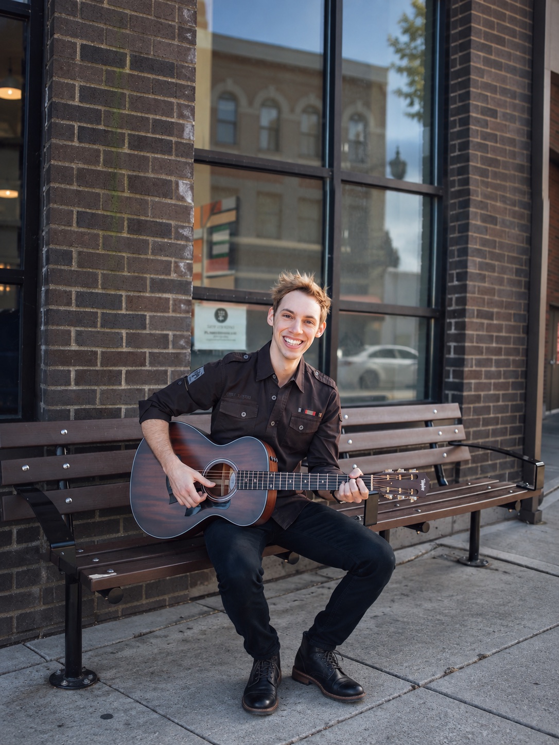 Derik Nelson smiles while playing an acoustic guitar on a bench.
