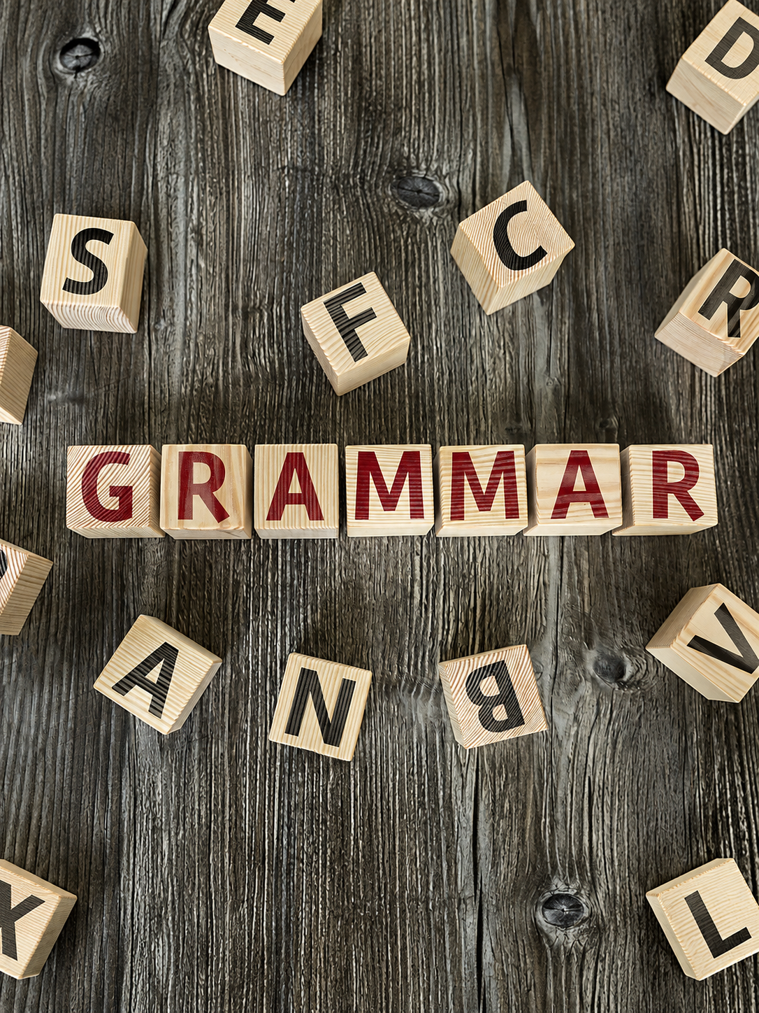 Wooden letter tiles spell “grammar” on a dark wood surface surrounded by scattered alphabet blocks.