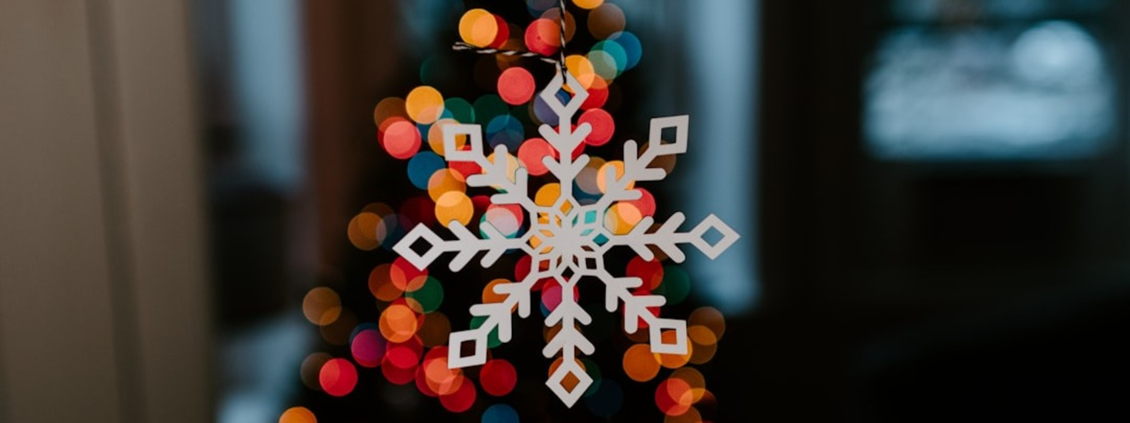 A white snowflake ornament hangs in front of a Christmas tree with colorful lights.